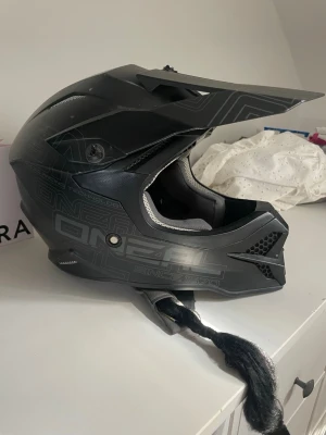Svart crosshjälm med fläta - Säljer en svart crosshjälm med grå detaljer och grafiskt mönster. Hjälmen har ett långt visir och en fastsatt svart fläta baktill. Invändigt är den vadderad med grått tyg för extra komfort. Perfekt för motocross eller downhill.