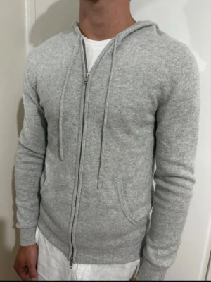 John henric zip hoddie - Säljer en ljusgrå hoodie med dragkedja och huva. Hoodien har dragsnören, ribbade muddar och fickor framtill. Tillverkad i ett mjukt, stickat material som känns riktigt skönt mot huden. Perfekt för dig som gillar en clean och enkel stil.