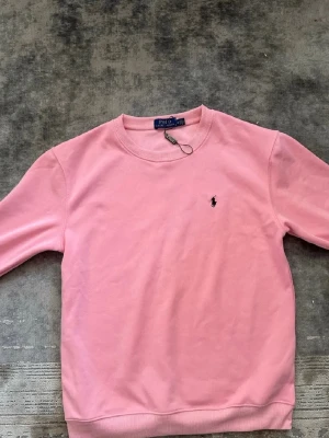 Rosa sweatshirt från Polo Ralph Lauren - En stilren rosa sweatshirt från Polo Ralph Lauren med rund halsringning och den klassiska lilla loggan broderad på bröstet. Tröjan har ribbade muddar vid ärmslut och nederkant. Perfekt för en avslappnad och trendig look.