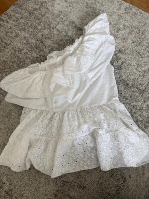 Vit oneshoulder klänning med volang - Söt vit oneshoulder klänning med volangdetaljer och broderad spets. Klänningen har en luftig passform och är perfekt för varma dagar. Materialet känns lätt och somrigt, och den har ett romantiskt uttryck med sina broderade detaljer.