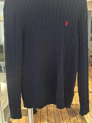 Mörkblå kabelstickad tröja Polo Ralph Lauren - Mörkblå kabelstickad tröja från Polo Ralph Lauren med klassisk röd logga broderad på bröstet. Tröjan har rund halsringning, långa ärmar och ribbade muddar vid ärmslut och nederkant. Perfekt för lager på lager och stilren look.
