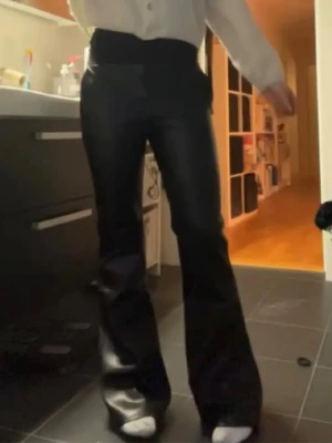 Svarta utsvängda byxor i fuskskinn - Säljer ett par svarta byxor i fuskskinn med hög midja och utsvängda ben. Snyggt glansigt material som ger en trendig look. Perfekta för dig som vill sticka ut lite extra och gillar en modern stil.