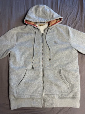 Grå zip hoodie från Burberry - Snygg grå hoodie med dragkedja från Burberry. Utsidan är enkel och stilren med två fickor och liten logga på bröstet. Insidan av huvan har det klassiska Burberry-rutiga mönstret i beige, svart och rött. Mjuk bomullskänsla och ribbade muddar.