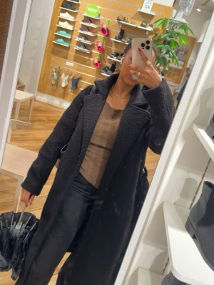 Lång svart ullkappa med krage - Snygg lång svart kappa i ullmix med bred krage och rak passform. Perfekt för kyliga dagar och ger en stilren look. Kappan har diskreta fickor i sidorna och stängs framtill. Passar till både jeans och finare outfits.
