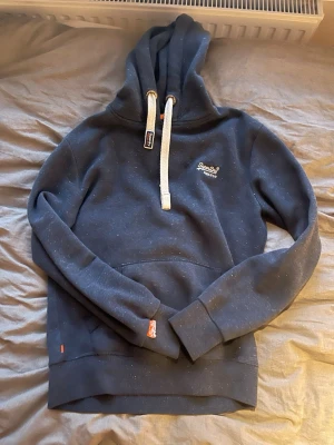 Blå hoodie från Superdry med huva - Mörkblå hoodie från Superdry med vita snören i huvan och broderad logga på bröstet. Hoodien har känguruficka framtill och ribbade muddar vid ärmslut och nederkant. Perfekt för en avslappnad och stilren look.