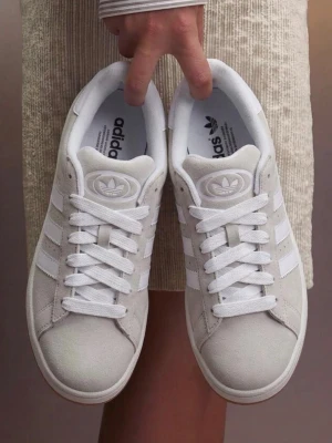 Vita Adidas sneakers i mocka - Snygga beigea Adidas sneakers i mocka med klassiska tre ränder på sidan och gummisula. Skorna har en låg profil och vita skosnören. Perfekta för dig som gillar stilrena och tidlösa sneakers med en sportig touch.