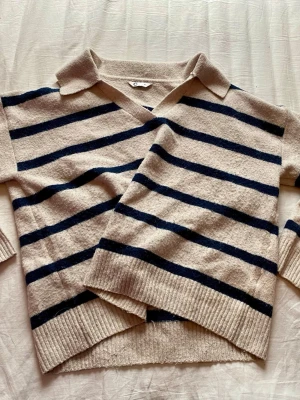 Beige stickad tröja med blå ränder - Stickad tröja i beige med mörkblå horisontella ränder och krage. Tröjan har v-ringning och ribbade muddar vid ärmslut och nederkant. Perfekt för dig som gillar en avslappnad men ändå stilren look.