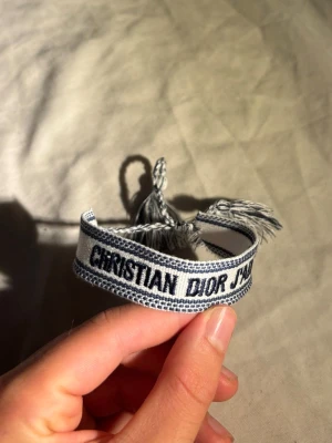 Dior tygarmband - Säljer ett snyggt tygarmband från Christian Dior med texten 'J'adior' i svart på vit bakgrund. Armbandet har knytband med tofsar i ändarna och är perfekt för dig som gillar exklusiva accessoarer med streetstyle-vibe.