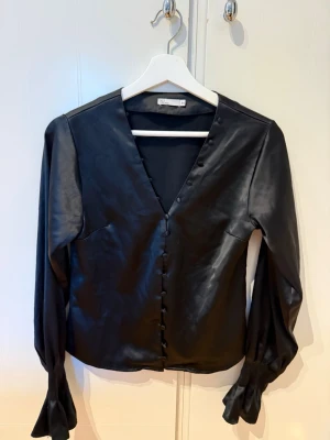 Svart satinblus med knappar - Stilren svart blus i satin med v-ringning och klädda knappar framtill. Blusen har långa ärmar med lätt puff och volang vid ärmslut. Perfekt för dig som gillar en elegant och trendig look.