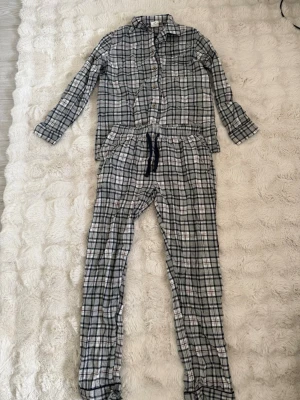 Rutiga pyjamasbyxor och skjorta S - Mysig pyjamas i två delar med rutigt mönster i grått, vitt, marinblått och vinrött. Skjortan har knappar framtill och klassisk krage, byxorna har dragsko i midjan. Perfekt för chillkvällar hemma.