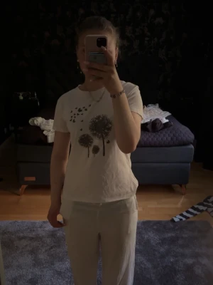 Vit t-shirt med maskrosmotiv - Säljer en vit t-shirt med tryck av maskrosor och flygande frön på framsidan. T-shirten har rund hals och korta ärmar. Perfekt för dig som gillar naturinspirerade prints och en enkel, clean stil.