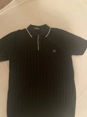 tshirt BOSS - Säljer en svart pikétröja från BOSS med diskreta vertikala ränder och vit piping på kragen. Tröjan har korta ärmar, knappar framtill och en liten BOSS-logga på bröstet. Perfekt för en clean och stilren look.