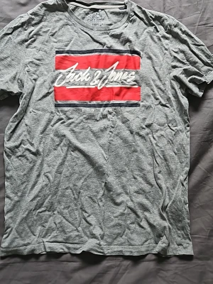Grå Jack & Jones t-shirt med tryck - Snygg grå t-shirt från Jack & Jones med korta ärmar och ett stort tryck i rött, vitt och blått på bröstet. Tillverkad i mjuk bomull och har en klassisk passform som funkar till det mesta. Perfekt för en avslappnad stil.