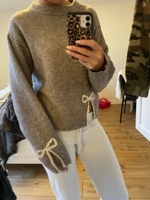 Beige stickad tröja  - Mysig beige grå stickad tröja. Tröjan har dekorativa vita spetsrosetter på ena ärmen och vid sidan av midjan. Tröjan är från zara i storlek s-xs.❤️