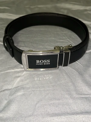Svart skärp från Hugo Boss - Snyggt svart skärp från Hugo Boss med stilren silverfärgad metallspänne och tydlig logga framtill. Skärpet är gjort i slätt skinn och har klassisk design som passar till många olika outfits. Priset kan diskuteras vid snabb affär.