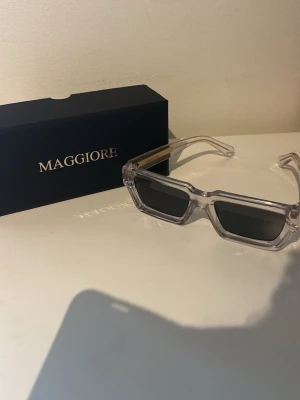Transparenta solglasögon Maggiore - Säljer ett par trendiga solglasögon från Maggiore i sprillans nytt skick med transparent båge och rektangulära svarta glas. Snygga gulddetaljer på skalmarna ger en extra touch. Perfekta för dig som vill sticka ut med en modern look.