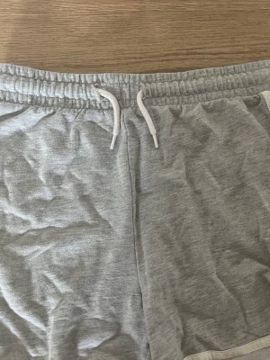 Grå mjukisshorts med snörning - Snygga och bekväma grå mjukisshorts med vit snörning i midjan och vita detaljer längs sidorna. Perfekta för chill dagar eller träning. Elastisk midja för skön passform.