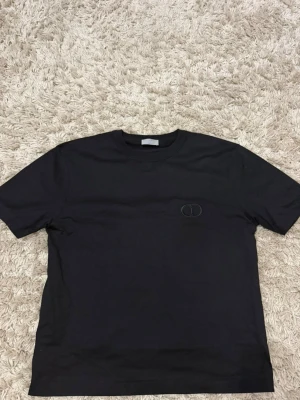 Svart basic t-shirt med diskret logga - En svart t-shirt i klassisk modell med korta ärmar och rund hals. T-shirten har en liten, diskret broderad logga på bröstet. Tillverkad i mjukt bomullsmaterial som känns skönt mot huden. Perfekt för en clean och stilren look.