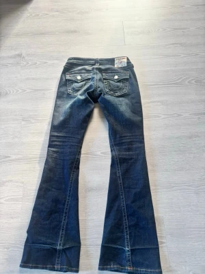 True religion Mörkblå bootcut jeans med fickdetaljer - Säljer ett par mörkblå bootcut jeans med slitningar och snygga detaljer på bakfickorna. Jeansen har dubbla knappar på fickorna bak och kontrastsömmar. Materialet är klassisk denim i bomull. Perfekta för dig som gillar retrovibbar och markerad siluett. Knappt använda 