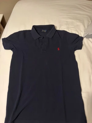 Mörkblå pikétröja från Polo Ralph Lauren - Klassisk mörkblå pikétröja från Polo Ralph Lauren med korta ärmar och krage. Tröjan har knappar framtill och den ikoniska röda logotypen broderad på bröstet. Klassiker från polo Ralph Lauren som jag säljer nu eftersom jag har växt ut ur den, vid funderingar hör av dig vid dms 💪