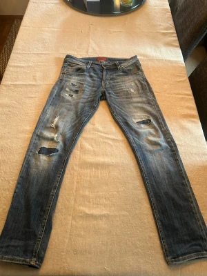 Jack and Jones jeans - Säljer ett par blå jeans med flera slitningar och hål på benen för en riktigt trendig och avslappnad look. Jeansen har klassisk femficksmodell och raka ben. Perfekta för dig som gillar streetstyle och vill ha ett par jeans med attityd.model:slim/glenn Storlek:W30L32