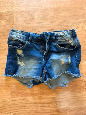 Blå denimshorts för flickor - Säljer ett par blå denimshorts med fransig kant nedtill. Perfekta för cool och fin stil i sommar. Strl 134