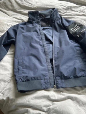  Peak Performance jacka - Snygg mörkblå vindjacka från Peak Performance med hög krage och dragkedja framtill. Jackan har ribbade muddar vid ärmslut och nederkant samt två sidofickor. Perfekt för blåsiga dagar och enkel att matcha med olika outfits.