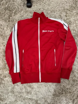 Röd Palm Angels zip-tröja - Snygg röd zip-tröja från Palm Angels med vita och svarta ränder längs ärmarna. Tröjan har hög krage, två fickor med dragkedja och ribbade muddar. Perfekt för en sportig och trendig look.