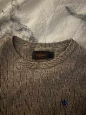 Morris KAN SÄNKA  - Säljer en beige kabelstickad tröja från Morris i 100% merinoull. Tröjan har rund halsringning, ribbade muddar och en liten blå broderad logga på bröstet. Perfekt för dig som gillar klassisk och stilren look.