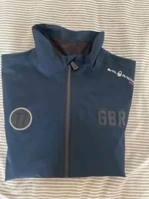 Marinblå vindjacka Sail Racing GBR - Snygg marinblå vindjacka från Sail Racing med hög krage, hel dragkedja och två fickor framtill. Jackan har tryckta detaljer som 'GBR' och '77', samt Sail Racing-logga på bröstet och ärmen. Materialet är tunt och vindtätt, perfekt för blåsiga dagar.