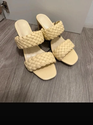 Gula flätade high heel sandaler - Säljer ett par trendiga gula sandaler med hög klack och två breda flätade remmar. Sandalerna har fyrkantig tå och är tillverkade i ett konstlädermaterial. Perfekta för dig som vill sticka ut med en snygg och modern look.