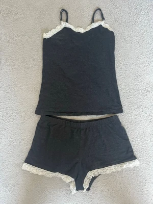 Mörkgrått spetskantat pyjamas-set - Säljer ett mörkgrått pyjamas-set med linne och shorts. Båda delarna har vita spetsdetaljer längs kanterna. Linnet har smala axelband och en enkel, rak passform. Shortsen har resår i midjan och matchande spets längs bensluten. Perfekt för mysiga kvällar. Skavanker/defekter finns men syns ej vid användning.