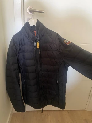 Svart dunjacka från Parajumpers - Säljer en äkta svart dunjacka från Parajumpers med quiltad design och hög krage. Jackan har dragkedja framtill, två sidofickor med dragkedja och en patch på ärmen. Insidan är grå med flera fickor och orange detaljer. Perfekt för kalla vinterdagar. Jackan är använd väldigt sparsamt och nypriset var 5000kr! Priset är förhandlingsbart!