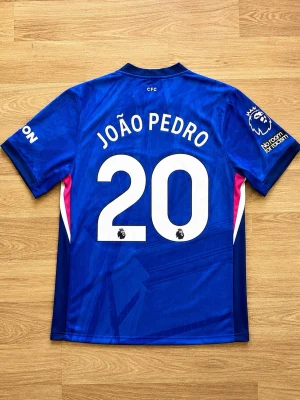 Chelsea FC matchtröja João Pedro - Säljer en blå Chelsea FC matchtröja från Nike med João Pedro och nummer 20 på ryggen. Tröjan har Premier League-logga på ärmen, klubbmärke och Nike-logga framtill samt vita och rosa detaljer på sidorna. Perfekt för fotbollsfans!