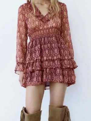 Kort blommig volangklänning från Zara - Supersöt kort klänning med blommigt mönster i vinröda och rosa toner. Klänningen har långa ärmar, smock i midjan och flera volanglager i kjolen. Tillverkad i ett lätt och luftigt material, perfekt för varmare dagar.