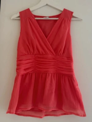Kort rosa omlotttopp - Snygg kort rosa klänning med omlottdesign och markerad midja. Klänningen är ärmlös och har ett lätt, luftigt tyg som ger en avslappnad känsla. Perfekt för varma dagar och somriga tillfällen.