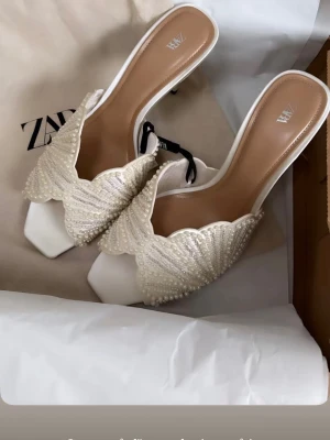 Vita pärlprydda pumps från Zara - Säljer ett par vita pumps från Zara med öppen häl och fyrkantig tå. Skorna har ett elegant pärlbroderi över hela ovandelen och en unik vågig kant. Perfekta för dig som vill sticka ut med en chic och trendig look. SKRIV VID INTRESSE OCH BUDA OM PRIS! 