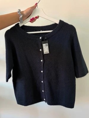 Mörkblå kofta med pärlknappar - Kortärmad mörkblå kofta med rund hals och pärlknappar framtill. Koftan har en mjuk, lite luddig yta och en enkel, stilren design som passar perfekt till jeans eller kjol.