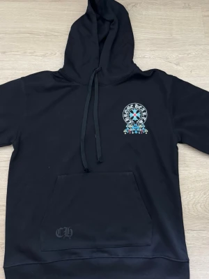 Svart Chrome Hearts hoodie med tryck - Svart hoodie från Chrome Hearts med stor färgglad kors- och logotyptryck på ryggen och mindre tryck på bröstet. Klassisk känguruficka framtill och justerbar huva med snören. Materialet känns mjukt och bekvämt, perfekt för en streetwear-look.