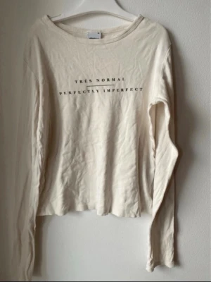 Vit långärmad topp från Gina Tricot - Vit långärmad topp från Gina Tricot med trycket 'TRÈS NORMAL PERFECTLY IMPERFECT' på bröstet. Tillverkad i mjuk bomull och har en avslappnad passform med rund halsringning. Perfekt för en chill och enkel stil.