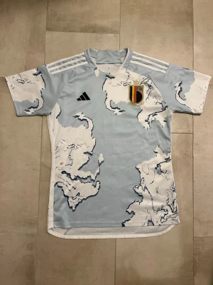 Belgien Adidas matchtröja ljusblå - Säljer en ljusblå matchtröja från Adidas med Belgiens landslagsemblem på bröstet. Tröjan har ett unikt mönster i vitt och mörkblått, samt klassiska Adidas-ränder på axlarna. Perfekt för fotbollsträning eller att bära till vardags.
