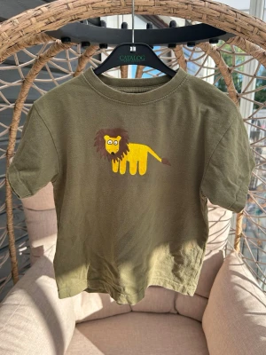 T-shirt strl. 100 - Säljer en grön t-shirt från Novita med ett gulligt lejontryck på bröstet. T-shirten har korta ärmar och är gjord i mjuk bomull, perfekt för en lekfull stil. 