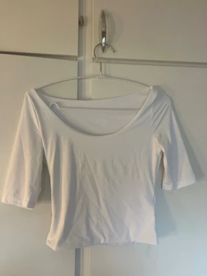 Vit topp med trekvartsärm - Säljer en enkel och stilren vit topp med trekvartsärm och bred urringning. Toppen har en tight passform och är tillverkad i ett mjukt, stretchigt material som sitter snyggt på kroppen. Perfekt att matcha med jeans eller kjol.