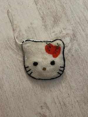 Hello Kitty pärlplånbok med jordgubbe - Söt liten plånbok formad som Hello Kittys ansikte, täckt av vita och svarta pärlor med en röd jordgubbe som detalj. Har en praktisk handledsrem och är perfekt för småsaker. Plånboken är unik och sticker ut med sitt lekfulla utseende.