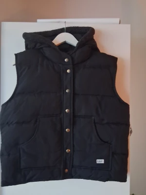 Svart dunväst med huva. Lee Storlek Large  - Snygg svart dunväst med stor huva och tryckknappar framtill. Västen har två stora fickor och ett mjukt foder i huvan. Perfekt för lager på lager och håller dig varm under kyliga dagar.
