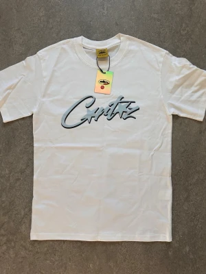 Vit t-shirt Corteiz - Snygg vit t-shirt från Critiz med stort svart och grått tryck på bröstet. Klassisk passform med rund hals och korta ärmar. Perfekt till jeans eller shorts för en clean och stilren look. Perfekt för de som har size S.