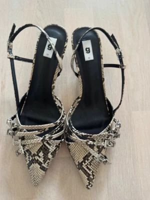 Snake print heels - Snygga och  med spetsig tå och ormmönstrat material i beige och svart. Skorna har flera tunna remmar med små spännen och en elegant klack. Perfekta för dig som vill sticka ut med en trendig och edgy look. Snake print
