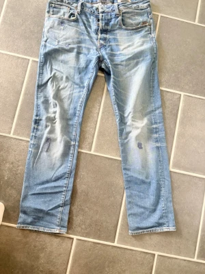 Ljusblå raka jeans med slitningar - Säljer ett par ljusblå jeans med raka ben och slitna detaljer på både fram- och baksida. Jeansen har klassisk femficksdesign och tydliga tvättade partier för en avslappnad look. Perfekta för dig som gillar en casual och trendig stil.