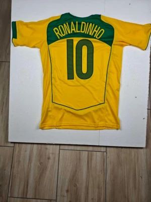 Gul Brasilien fotbollströja Ronaldinho - Säljer en ikonisk gul Brasilien fotbollströja från Nike med gröna detaljer, nummer 10 och Ronaldinho på ryggen. Tröjan har rund hals, korta ärmar och är tillverkad i lätt och ventilerande material. Perfekt för dig som älskar fotboll och vill sticka ut på planen.