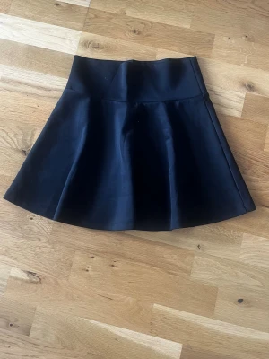 Svart kortkjol med utsvängd form - Säljer en svart kortkjol med hög midja och utsvängd siluett. Kjolen har en enkel och stilren design som passar till många olika looks. Perfekt för dig som gillar en klassisk men trendig stil.